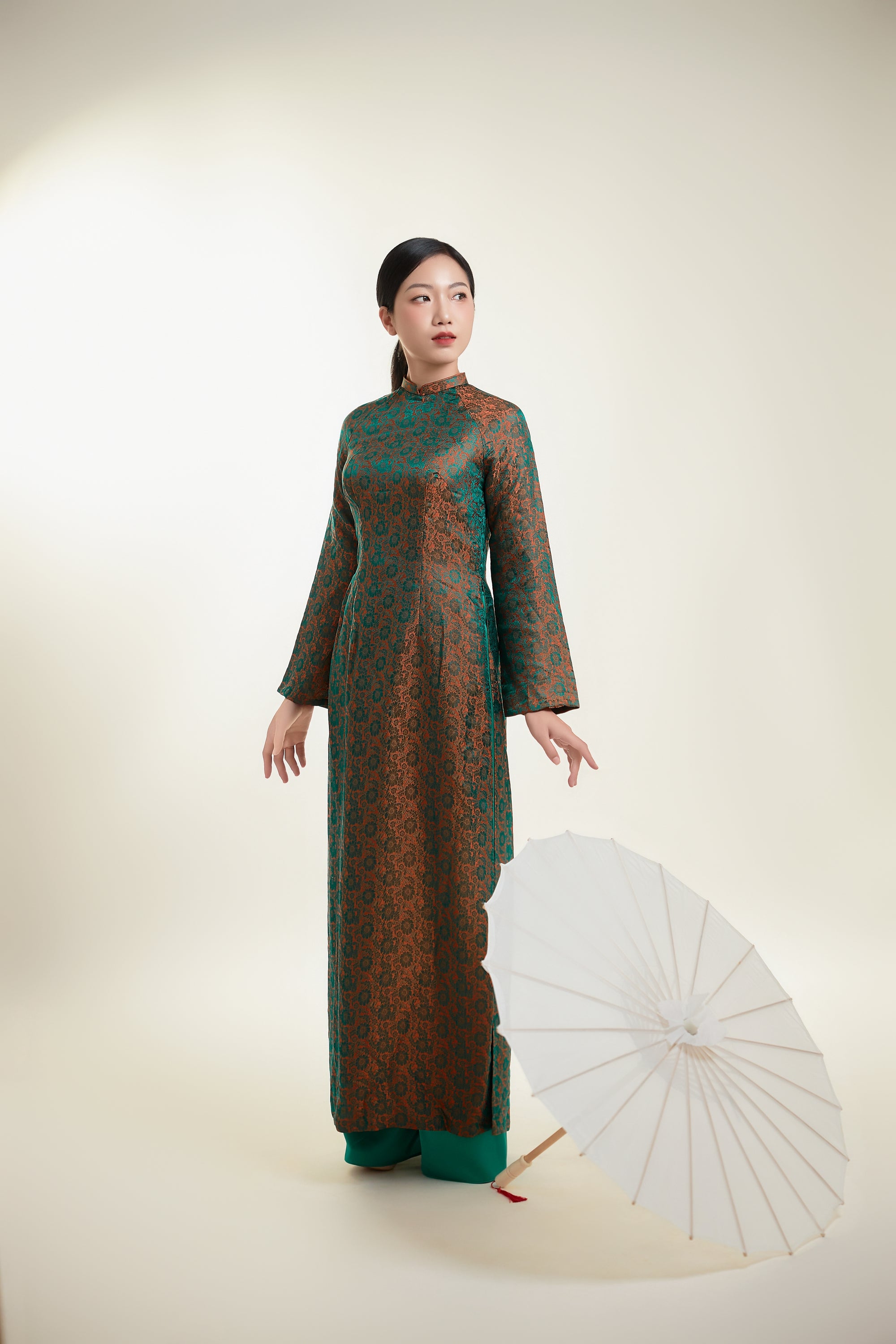 Elegant 100% silk Ao Dai in green and gold with intricate patterns, paired with a white parasol.