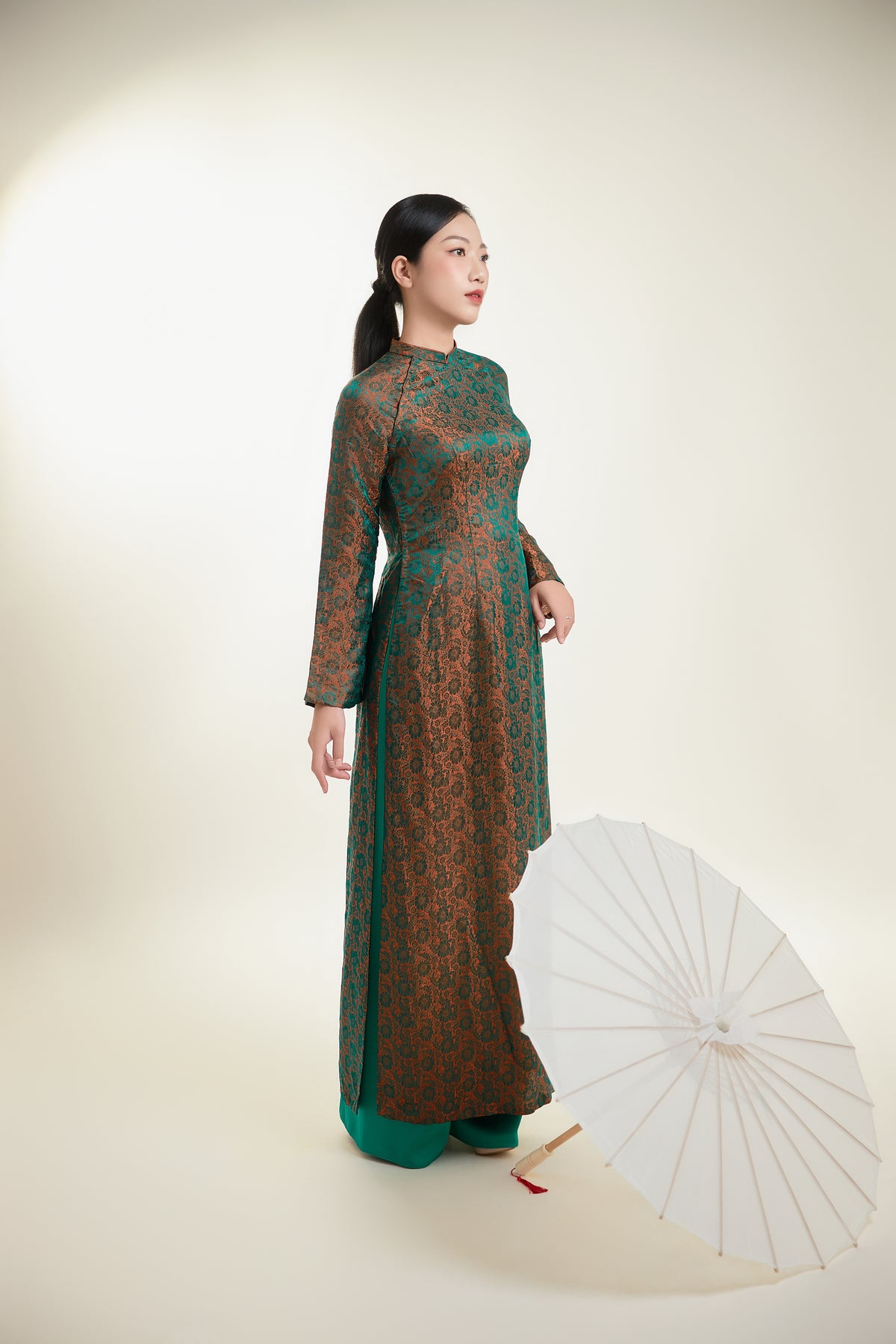 Full view of a green and gold silk Ao Dai with a white parasol accentuating its elegance.